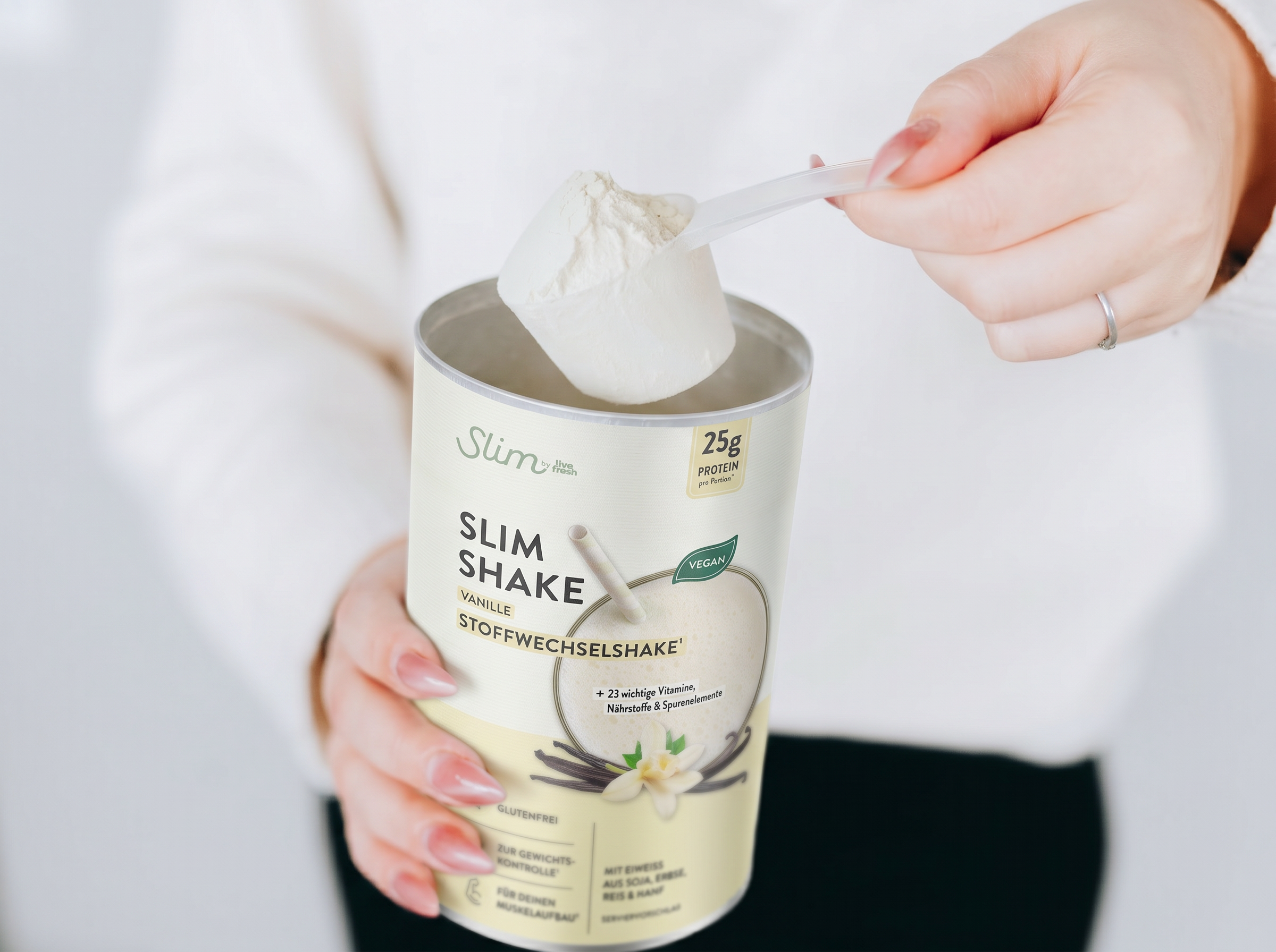 Hand hält einen Messlöffel mit Pulver über einer Dose „Slim Shake Vanille Stoffwechselshake“, die für Gewichtsreduktion beworben wird. Der Stoffwechselshake wird portioniert, um beim Abnehmen zu unterstützen, während Angaben wie „25g Protein“ und „vegan“ auf der Verpackung sichtbar sind.